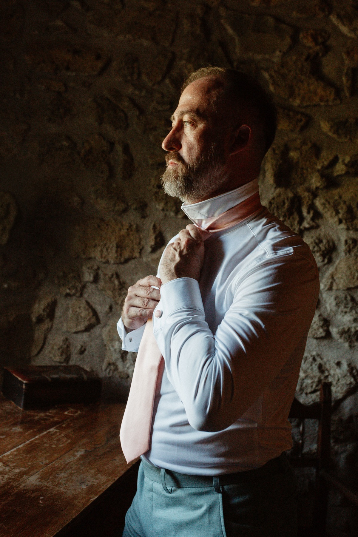 Ken against a stone wall, knotting his peach tie before the ceremony